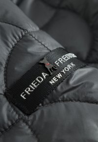 Gequilte zwarte nylon stof met een glanzende textuur, voorzien van een merklabel in het zwart met een zilveren logo en een rood accent op het kraaggebied.