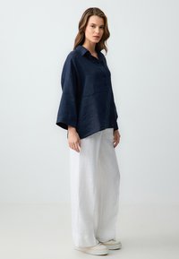 Chemise en lin navy avec un col, trois boutons et des manches larges, associée à un pantalon large en lin blanc et des chaussures décontractées.