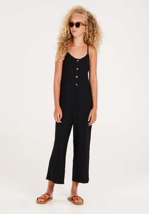 Combinaison noire côtelée avec un corsage ajusté et un pantalon à jambes larges, présentant des boutons sur le devant et des fines bretelles. Associée à des sandales marron et des lunettes de soleil.