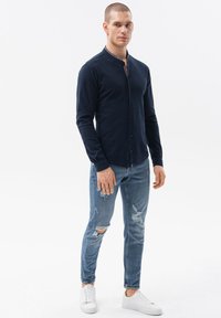 Tummansininen pitkähihaisten napitettava paita mandariinikauluksella, yhdistettynä vaaleansinisiin revittyihin farkkuihin ja valkoisiin lenkkareihin.