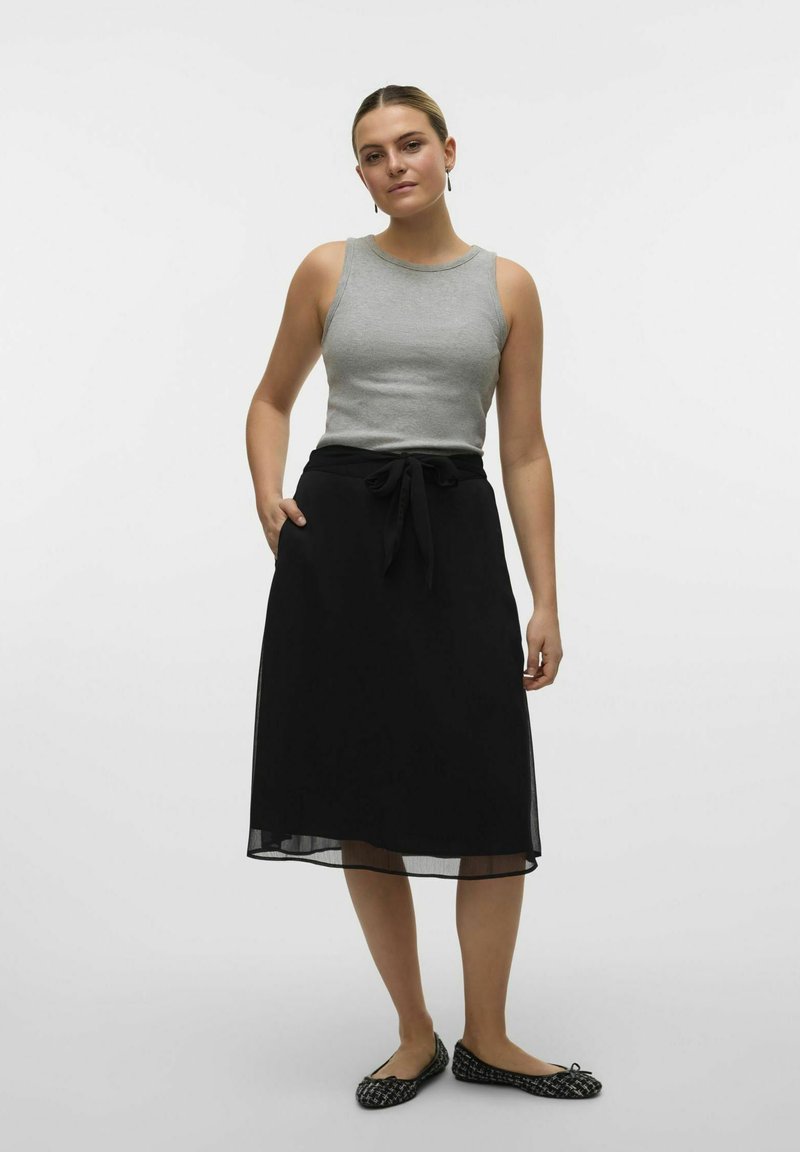 Top grigio senza maniche abbinato a una gonna nera con vita legata e tasche; scarpe nere flat con motivo completano l'outfit.