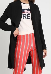 Černý kardigan s otevřeným předním dílem z měkké tkaniny, vrstvený design sahající přes světle zbarvené tričko s grafikou a červené pruhované legíny.