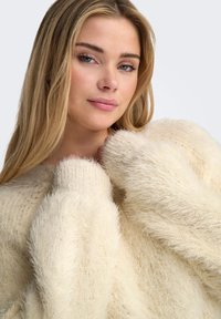 Cream-colored fuzzy sweater with a loose fit and ribbed cuffs. The texture is soft with a light, airy appearance.