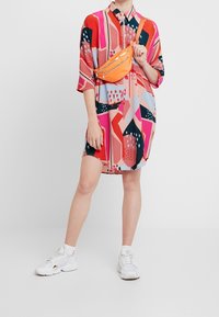 Robe chemise surdimensionnée colorée avec des motifs géométriques en rose, rouge et bleu. Assortie à une ceinture-sac orange vif et des baskets blanches.
