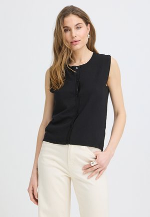 Femme aux longs cheveux châtain clair, portant un haut noir sans manches boutonné et un pantalon blanc cassé, debout avec une main dans la poche.