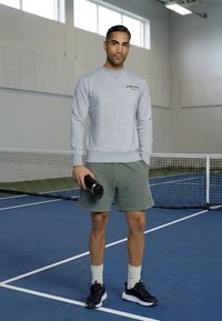 Mann i grå genser og grønne shorts som holder en svart vannflaske og står på en innendørs tennisbane.