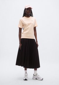 Ferskenfarvet T-shirt, sort plisseret nederdel, hvide sneakers og en pink kasket. T-shirten har et lille logo, og nederdelen har lodrette plisseringer for tekstur.