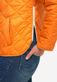 Giacca arancione trapuntata con finitura liscia, dotata di una cerniera bianca e un orlo arrotondato. Abbinata a jeans in denim blu.