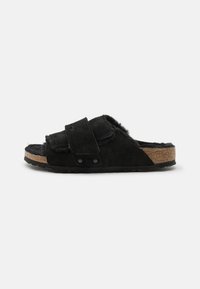 Black suede slide sandal with adjustable strap, soft fur lining, cork footbed, and textured rubber sole. Accented with black hardware.
