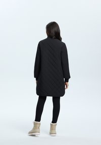 Manteau long noir matelassé avec col montant, manches longues et poignets côtelés, associé à des bottes d'hiver beiges en laine de mouton.