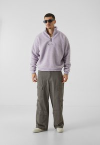 Pullover in pile di lavanda con zip a un quarto, caratterizzato da un piccolo logo. Abbinato a pantaloni cargo grigi a vestibilità ampia e sneakers bianche.