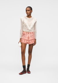 Femme portant un haut à manches longues en dentelle blanche avec des volants, un short rose à motifs, des chaussettes noires, des chaussures rouges, tenant une petite pochette blanche.