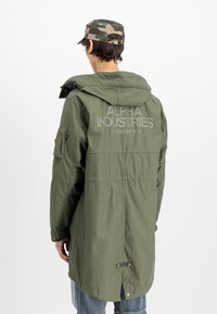 Άτομο που φοράει μακρύ πάρκα ελιές με το κείμενο "Alpha Industries Studio Edition" στην πλάτη και καπέλο παραλλαγής, με την πλάτη στραμμένη.