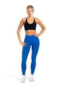 Czarny stanik sportowy i niebieskie legginsy z wysokim stanem. Gładka struktura, dopasowany krój. Modelka ma na sobie białe buty sportowe, ręce na biodrach.