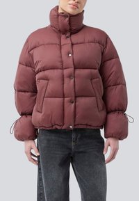 Person, der eine burgunderfarbene Steppjacke mit hohem Kragen und Druckknöpfen trägt, kombiniert mit dunkelgrauen Jeans, vor einem schlichten Hintergrund.