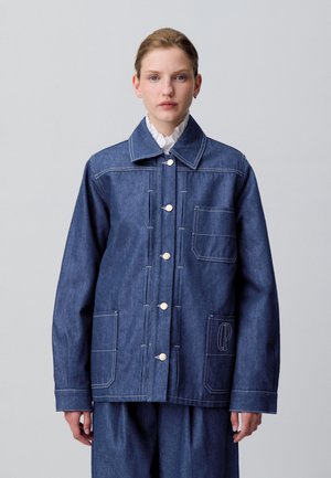 Jeune femme portant une veste en denim bleu avec de grandes poches et une blouse blanche à col volanté, se tenant devant un fond clair.