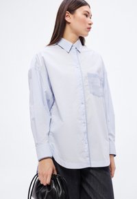 Svetlomodrá košeľa na zapínanie s náprsným vreckom, voľný strih, dlhé rukávy a jemná textura, doplnená o čiernu tašku.