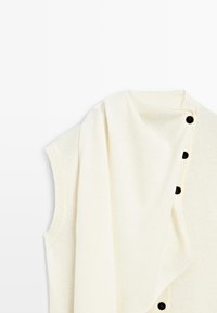 Cremfarbener, ärmelloser Top mit schwarzen Knöpfen, ausgestattet mit einem drapierten, weichen Stoffdetail an der Seite. Glatte Textur und schlichtes Design.
