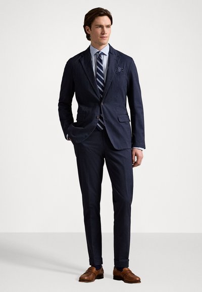 Blazer bleu marine à deux boutons, avec pantalon assorti et chemise habillée bleu clair. Comprend une cravate rayée et un mouchoir de poche à motifs. Chaussures en cuir marron.