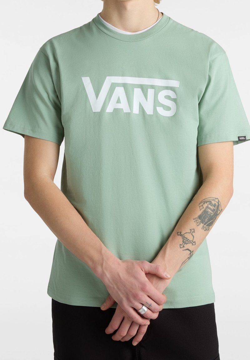 Personne portant un t-shirt Vans vert clair avec un logo blanc, un pantalon noir, des tatouages sur les bras et les mains jointes devant.