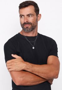 Zwart T-shirt van zachte stof, met meerdere zilveren kettingen, waaronder een ronde hanger. Model heeft een lichte baard en heeft de armen gekruist.