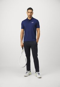 Ναυτικό μπλουζάκι πόλο με κοντά μανίκια και λογότυπο, συνδυασμένο με σκούρα παντελόνια και λευκά αθλητικά παπούτσια, κρατώντας μια ρακέτα τένις. Απλό φόντο.