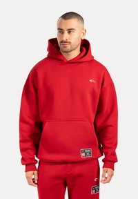 Červená mikina s kapucí z pevného materiálu, s klokankou, žebrovanými manžetami a logem a štítky značky na spodním lemu.