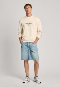 Felpa crema con testo stampato, abbinata a pantaloni shorts in denim blu chiaro con orlo sfrangiato e dettagli usurati, indossata con sneaker grigie e bianche.