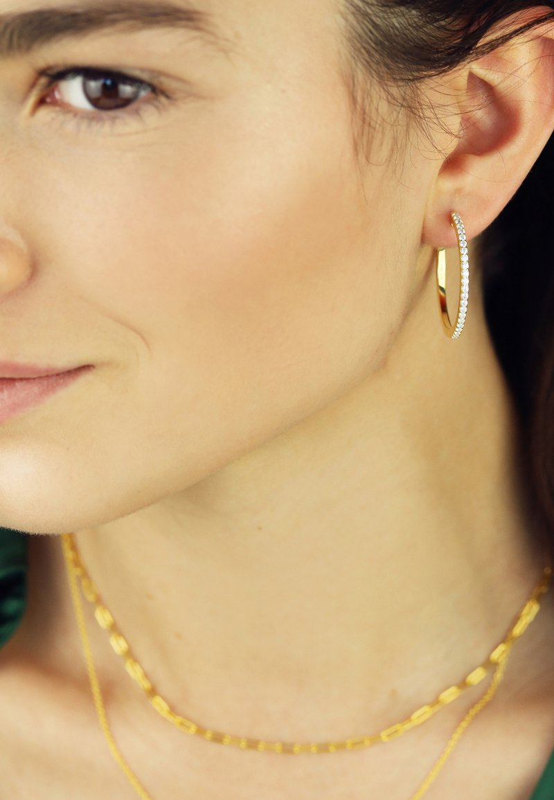 Goldene Creole mit einer Reihe klarer Kristalle, kombiniert mit geschichteten goldenen Ketten. Glatte Oberfläche und glänzende Ausführung.