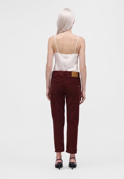 Femme aux longs cheveux blonds portant un top caraco en satin blanc, un pantalon bordeaux et des sandales noires à talons hauts, debout, dos face à l'avant.