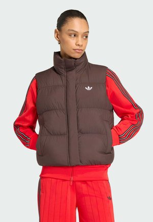 Donna con un gilet imbottito marrone sopra una tuta rossa Adidas con strisce nere, guarda verso sinistra con le mani nelle tasche del gilet.