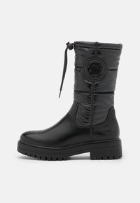 Botas negras hasta la rodilla con una parte superior brillante y acolchada, un detalle de logo redondo y una suela de goma texturizada para mayor agarre. Presenta un cierre con cordón.