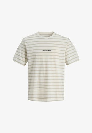 Uśmiechnięty chłopiec stojący, ubrany w jasną koszulkę w paski, luźne wyblakłe niebieskie dżinsy oraz czarne trampki z białymi podeszwami na jednolitym tle.