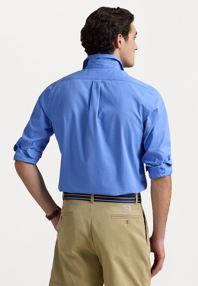 Camicia di cotone azzurro chiaro con maniche arrotolate, caratterizzata da una piega sul retro. Abbinata a pantaloni khaki e una cintura a righe.