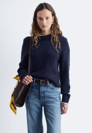 Jeune femme aux cheveux ondulés portant un pull bleu foncé et un jean bleu, tenant un sac à bandoulière sombre avec une écharpe jaune attachée.