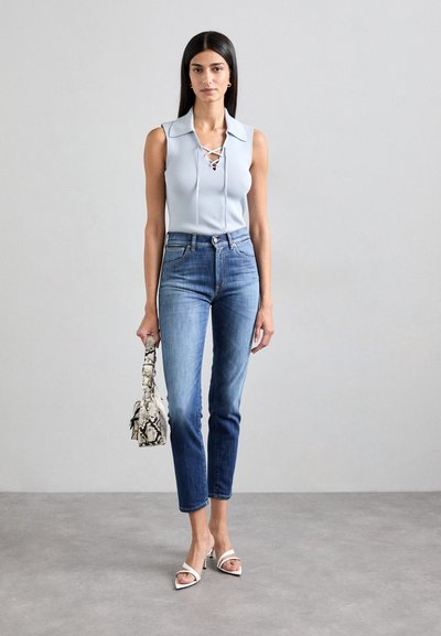 Top sin mangas de color azul claro con cuello y detalle de cordones, combinado con jeans de mezclilla azul, sandalias blancas de tacón y un bolso de mano con estampado de serpiente.
