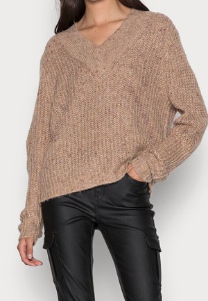 Femme portant un pull en maille beige ample avec un col en V et un pantalon cargo en cuir noir, main dans la poche, sur un fond uni.