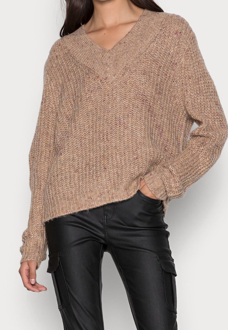 Femme portant un pull en maille beige ample avec un col en V et un pantalon cargo en cuir noir, main dans la poche, sur un fond uni.