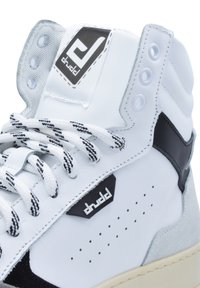 Zapatilla alta de cuero blanco con acentos en negro, que presenta paneles texturizados en gris, cordones con un patrón en blanco y negro, y una etiqueta con logo.