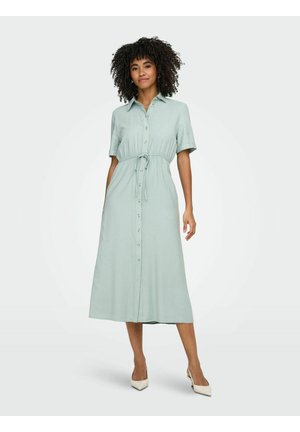 Nainen, jolla on kiharat hiukset, pukeutuneena vaaleanvihreään, lyhythihaiseen, napilliseen midi-mekkoon, jossa on vyötärönauha, seisomassa valkoisissa avokkaissa valkoista taustaa vasten.