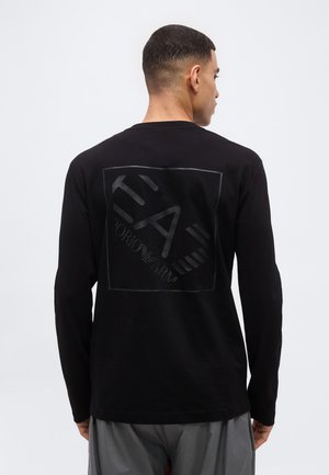 Άνδρας που κοιτάει προς τα πίσω, φοράει μαύρο μακρυμάνικο πουκάμισο με σκούρο λογότυπο Giorgio Armani τυπωμένο στο πίσω μέρος και γκρι παντελόνια.