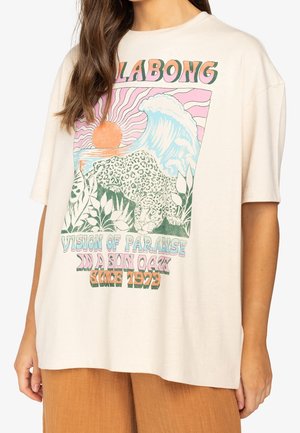 Kobieta nosząca beżową, oversize'ową koszulkę z kolorową grafiką lamparta, fali, słońca oraz napisem „Billabong Vision of Paradise in a Son OaK Since 1973".