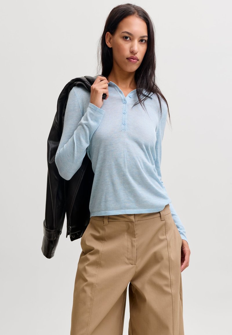 Femme portant une chemise bleu clair à manches longues et un pantalon beige, tenant une veste en cuir noire sur son épaule sur un fond uni.