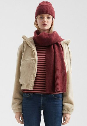 Beige Fleecejacke mit Seitentasche, bordeauxrot gestreiftes Oberteil und langer, strukturierter bordeauxroter Schal. Inklusive runder Brille und Strickmütze.