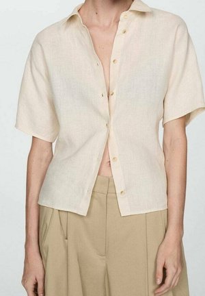 Szczupły tors w krótkim rękawie, jasnobeżowej koszuli na guziki częściowo rozpiętej, w połączeniu z luźnymi, wysokim stanem, beżowymi spodniami.