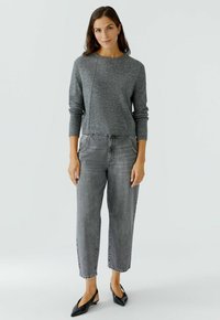 Frau in einem grauen Langarmpullover, hoch taillierten lockeren grauen Jeans und schwarzen spitzen Slingback-Schuhen, stehend vor einem weißen Hintergrund.