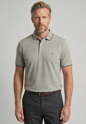 Mann mittleren Alters mit hellem Haar und Bart, der ein hellgraues Polohemd trägt, das in eine dunkelgraue Hose mit braunem Gürtel gesteckt ist.