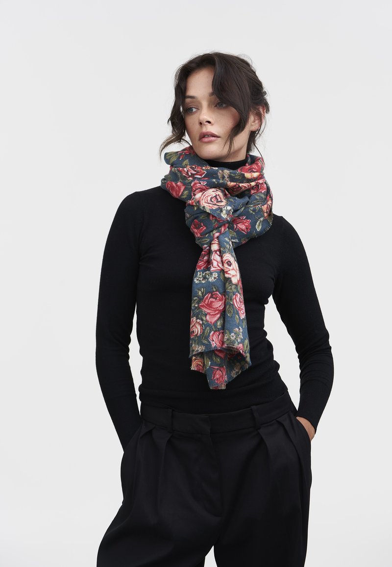 Mujer con cabello oscuro en un conjunto negro, llevando una bufanda floral azul alrededor del cuello, contra un fondo claro y uniforme.