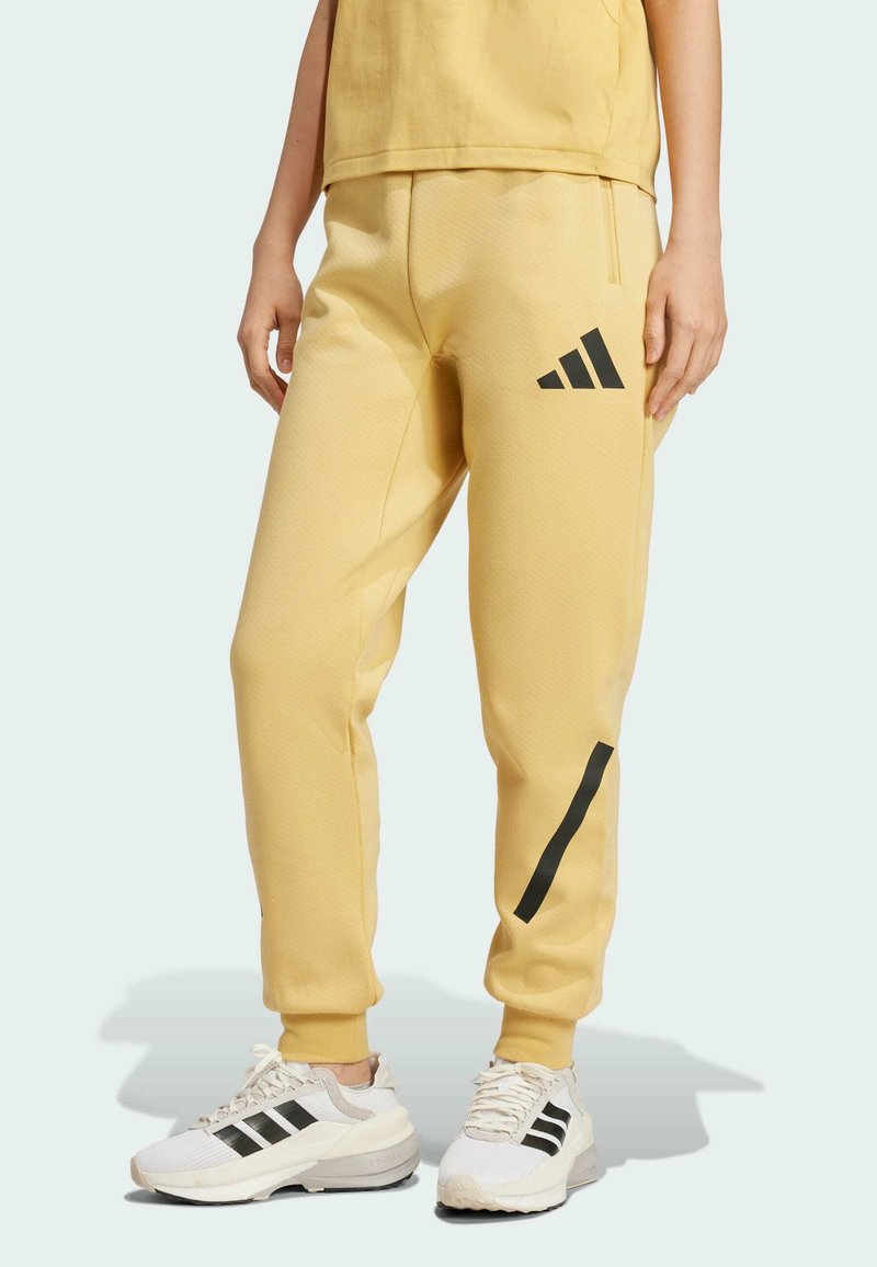 Persona che indossa pantaloni da jogging gialli con logo diagonale nero e scarpe da ginnastica bianche con strisce nere, in piedi su uno sfondo chiaro.