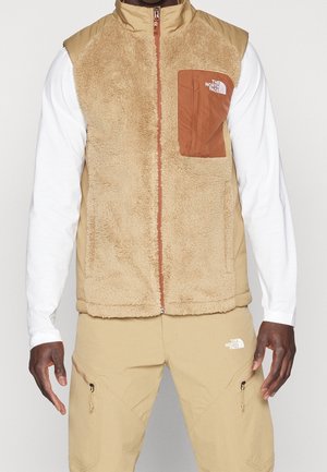 Uomo che indossa un gilet in pile beige con tasca sul petto marrone e logo, camicia a maniche lunghe bianca e pantaloni beige con tasche con cerniera.